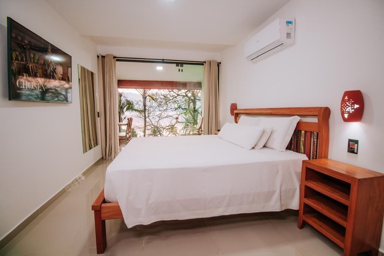 A hotel room with a bed, a wooden nightstand, a wall-mounted air conditioner, and a large window with curtains.