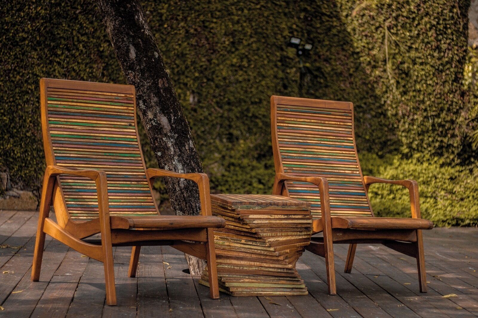 Dos sillones de madera con cojines a rayas, situados en una terraza de madera con un árbol y un seto al fondo.