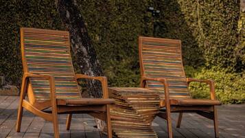 Dos sillones de madera con cojines a rayas, situados en una terraza de madera con un árbol y un seto al fondo.