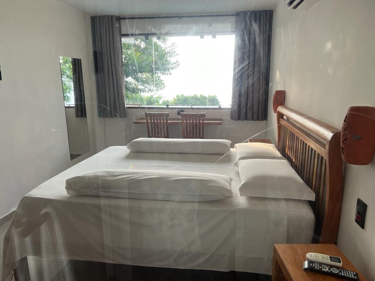 Habitación de hotel con cama, cabecero de madera, mesita de noche con control remoto y ventana con cortinas.