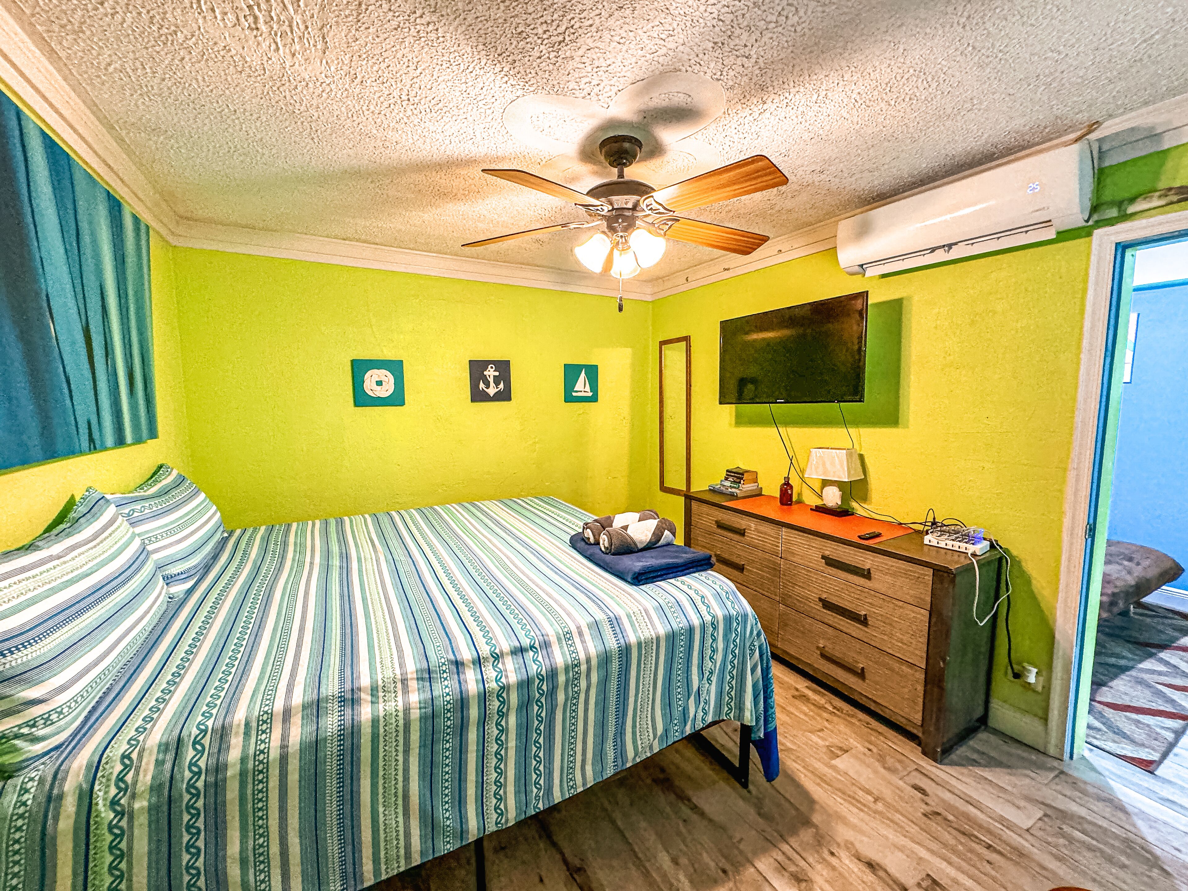 Une chambre comprenant un lit, une commode, un ventilateur de plafond, un téléviseur et un climatiseur fixé au mur.