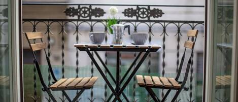 A balcony with a small table and two wooden chairs, a metal railing, and a view of a building in the background.