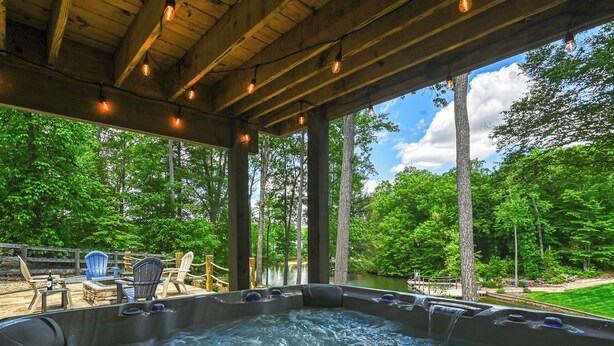 A hot tub area with wooden beams and string lights, surrounded by lush greenery and a view of a lake.