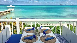 Una mesa con cuatro individuales tejidos y servilletas azules, con vista al mar turquesa y un pabellón blanco.