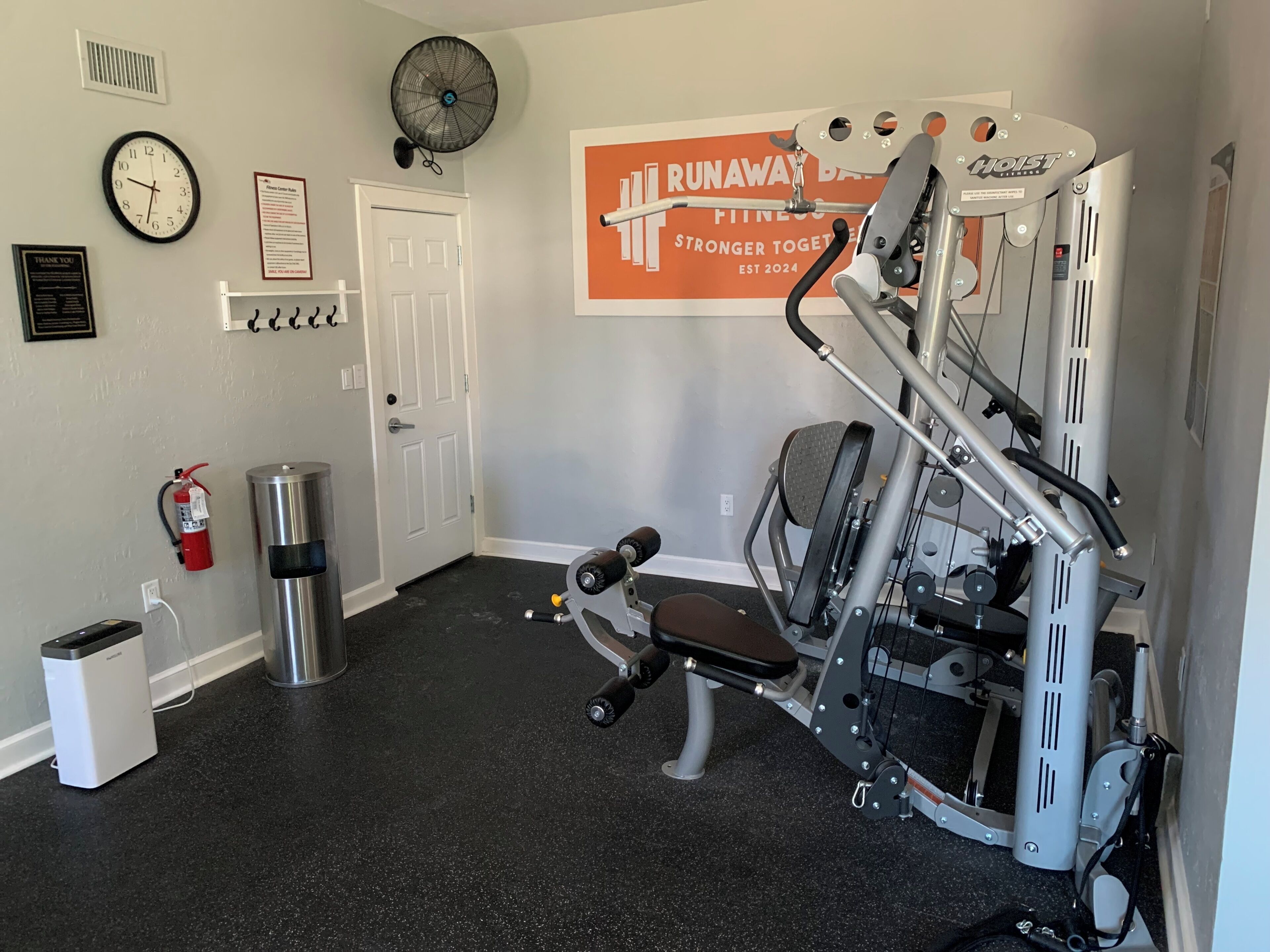 A gym with a wall-mounted fan, a clock, a fire extinguisher, a trash bin, and a 'Runaway Bay Fitness' sign.