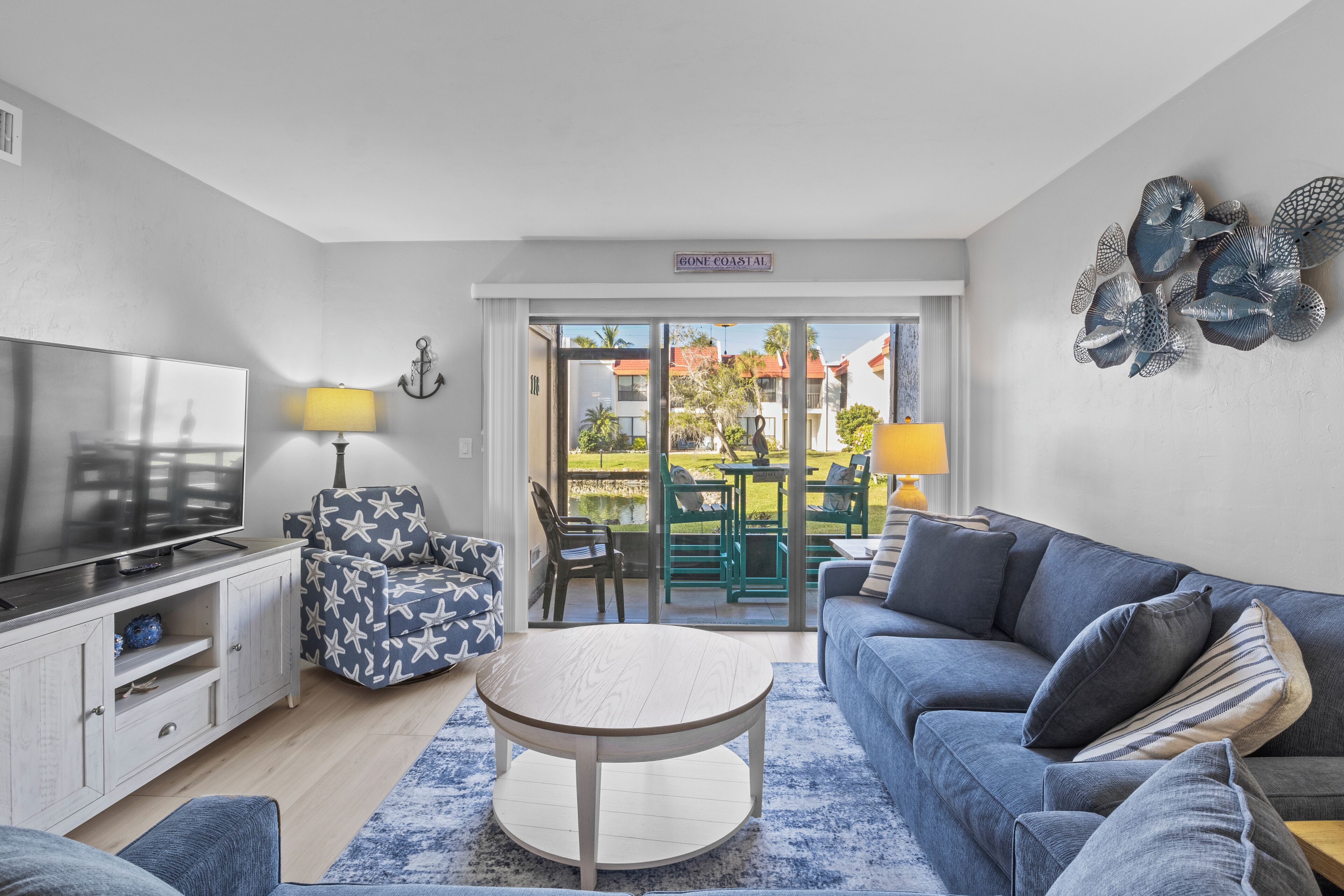 A modern living room with a flat-screen TV, a round coffee table, a blue sofa, and a patterned armchair. There is a sliding glass door leading to an outdoor area with a table and chairs.