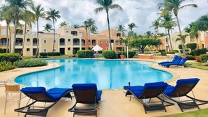Un área de piscina con sillones de descanso, palmeras y un edificio con balcones al fondo.