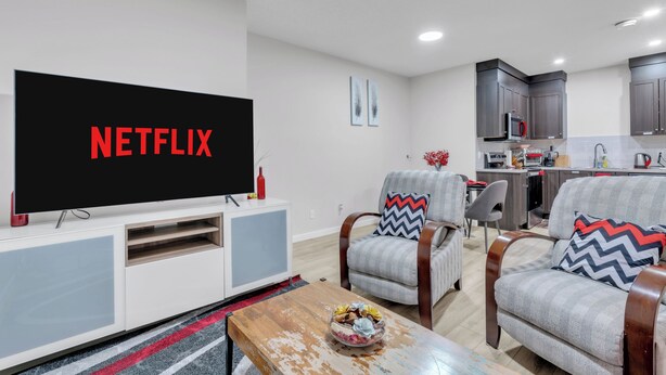 A modern living room with a flat-screen TV displaying "NETFLIX," two armchairs with patterned cushions, a wooden coffee table with a bowl, and a kitchen area in the background. - Beautiful Modern Unique Suite (Calgary)