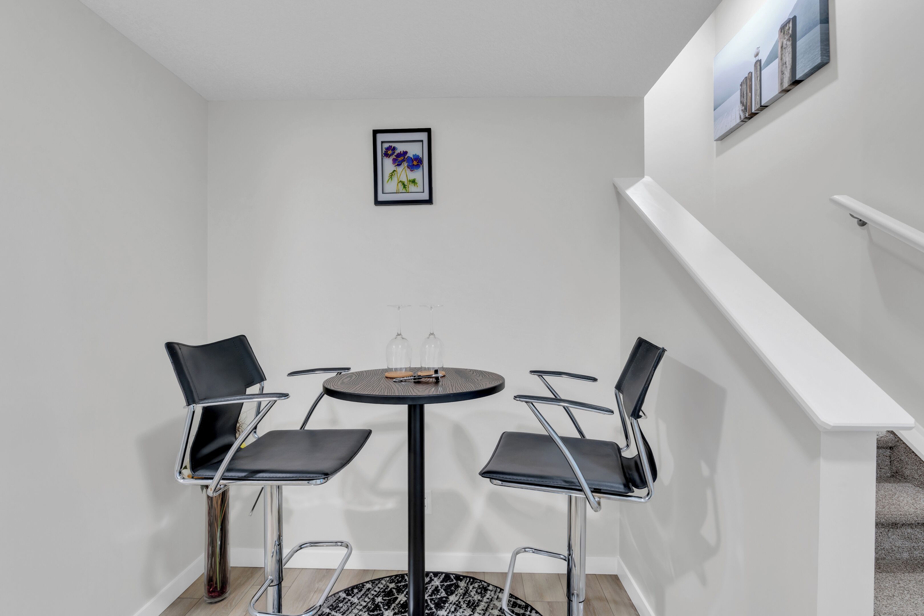 A corner room with a round table and four chairs, a framed picture on the wall, and a staircase to the right.