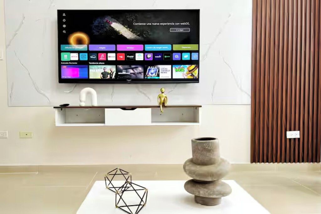 En moderne stue med et fladskærms-TV monteret på væggen, en minimalistisk hylde nedenunder og en dekorativ vase på et hvidt bord.