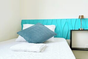Ein ordentlich bezogenes Bett mit einem weiß-blauen Kopfkissen, einem gefalteten weißen Handtuch und einem türkisfarbenen Kopfteil.