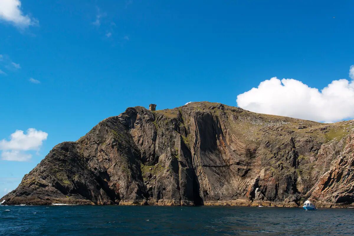 A rocky coastline with a small structure on top, a boat in the water, and a clear blue sky.