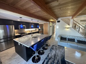 A modern kitchen with a central island, bar stools, and a dining area with a sofa. - Lakefront Blue House

License # STAR-04596 (Sylvan Lake)