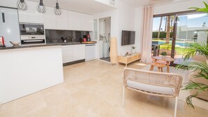 A modern kitchen with white cabinets, a Bosch oven, and a coffee maker. There is a wicker chair and a small table in the living area, with a TV mounted on the wall. - Marina Real apartment 504 (Manilva)