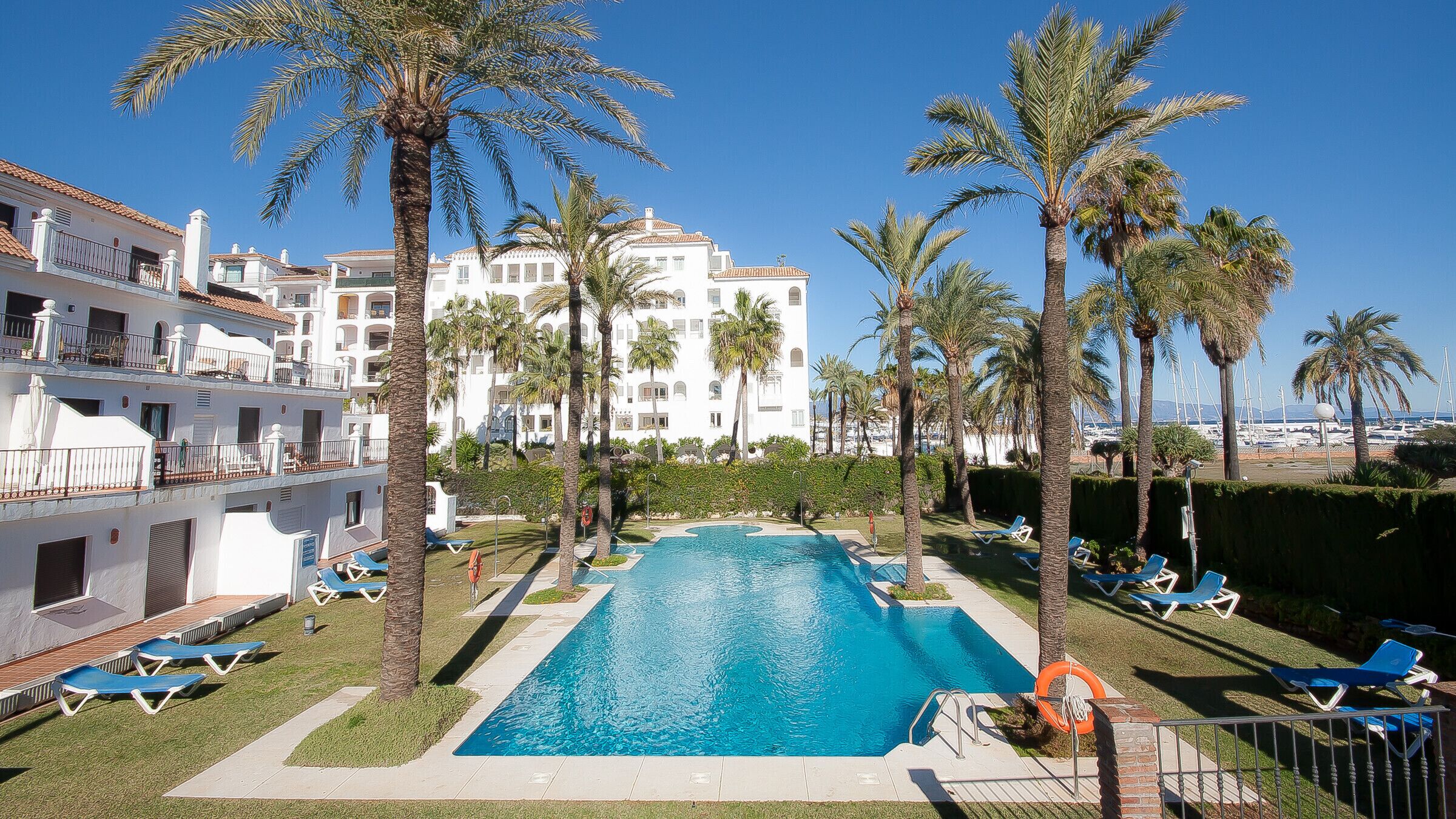 Una piscina rodeada de palmeras, con vista a un puerto deportivo y edificios blancos al fondo.