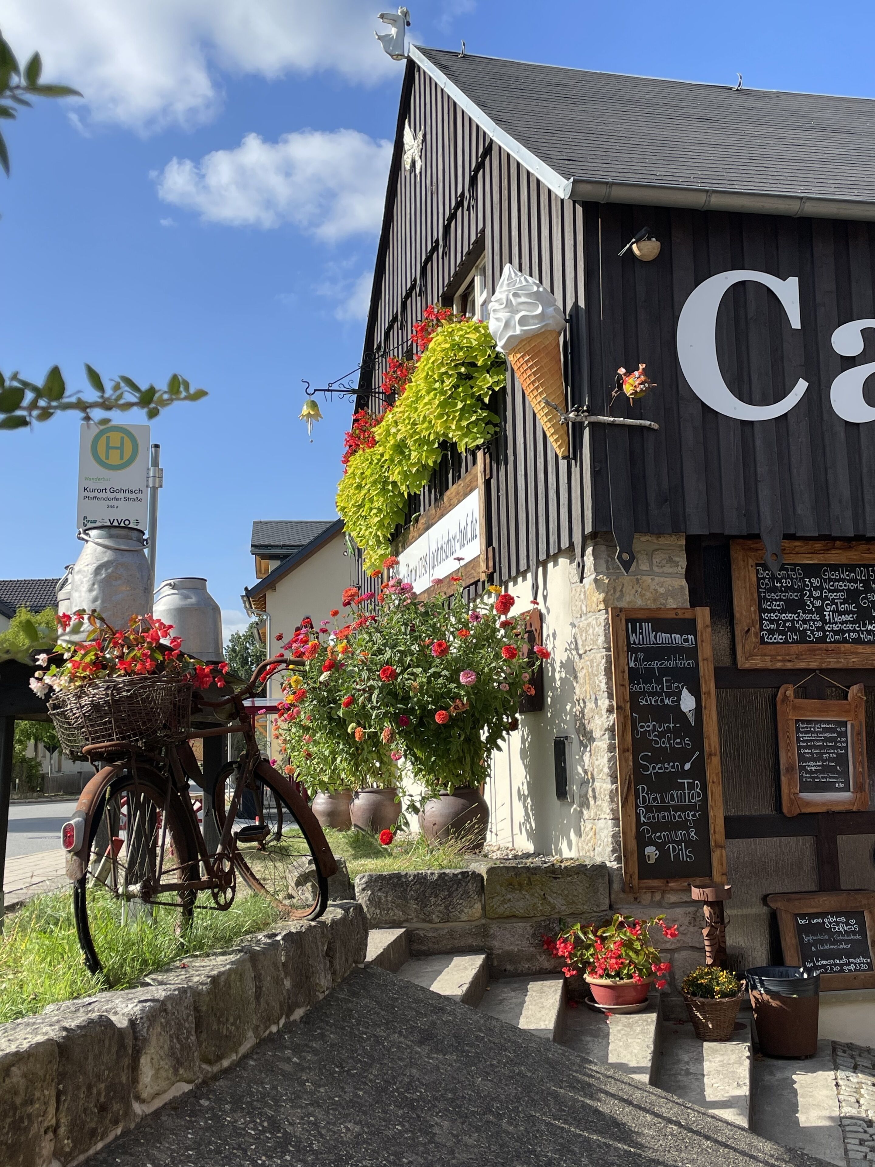 A building with a large sign reading "Café" and a signboard with a menu. There are flower baskets on the ground and a bicycle parked nearby.