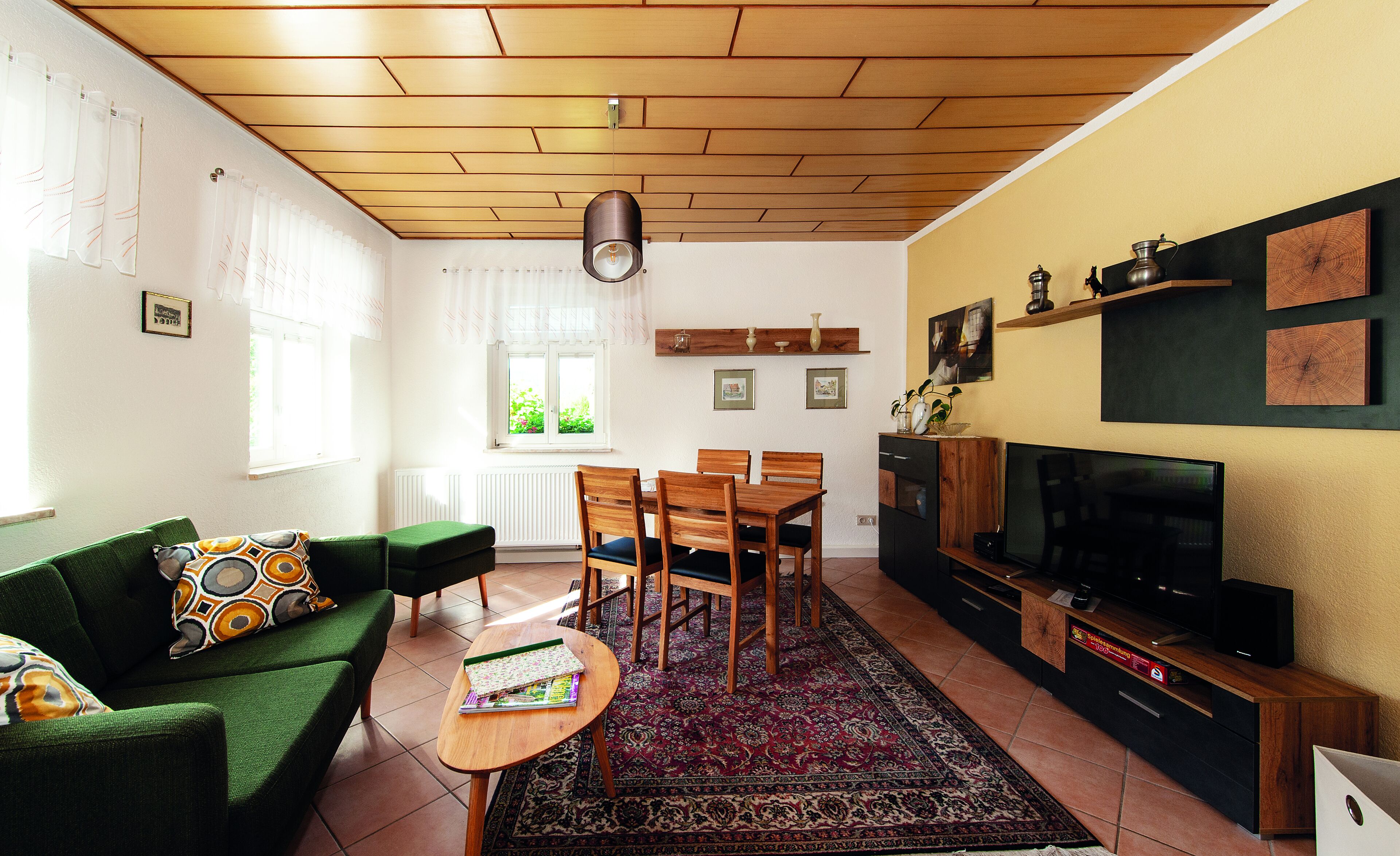 A living room with a green sofa, wooden dining table, and chairs, a TV, and a patterned rug.