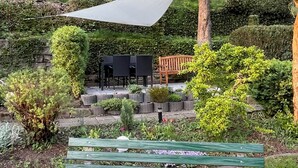 A garden with a wooden bench, a table, and chairs under a shaded area.