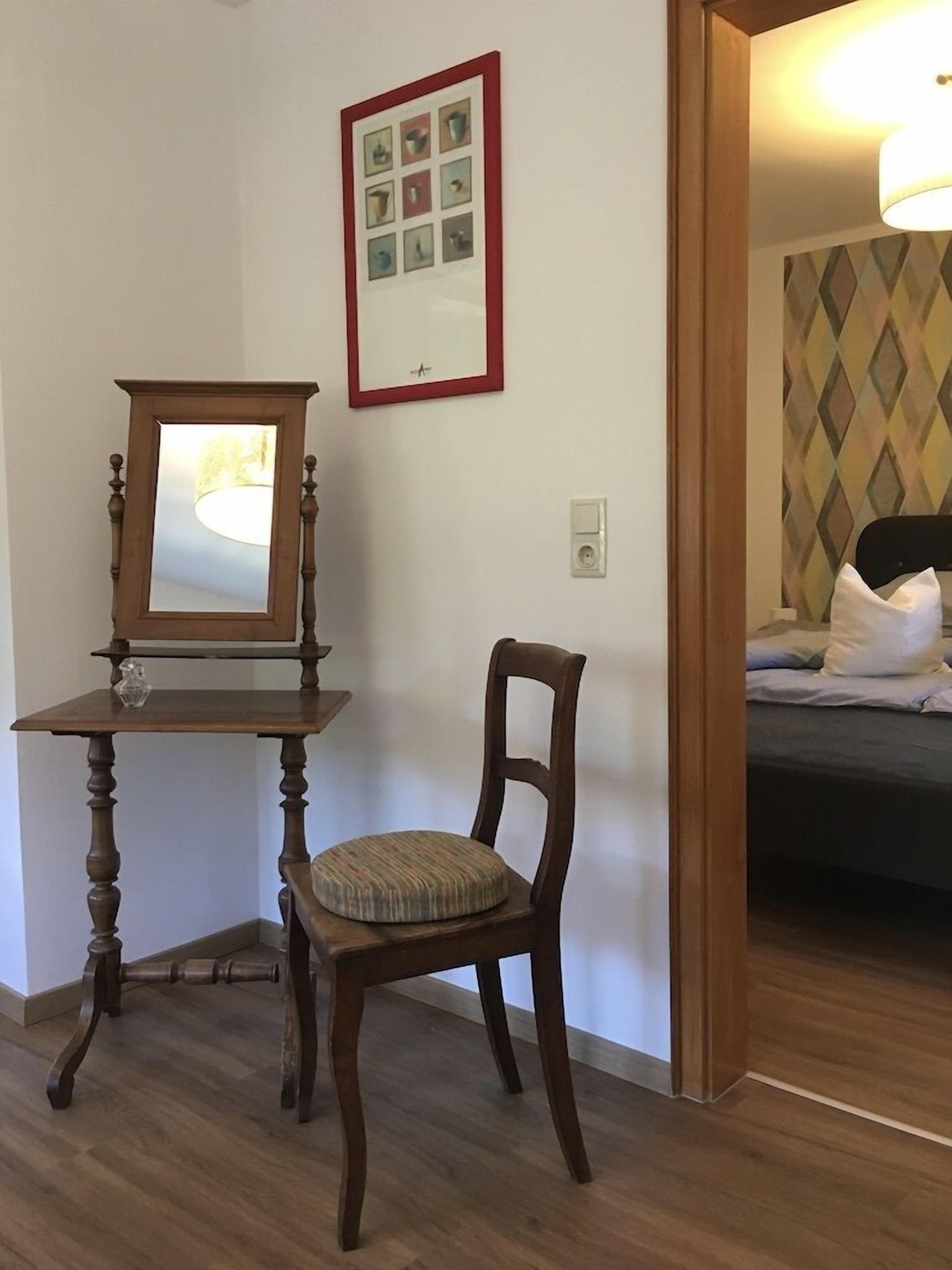 A room with a wooden chair, a small table with a mirror, and a bed with pillows.
