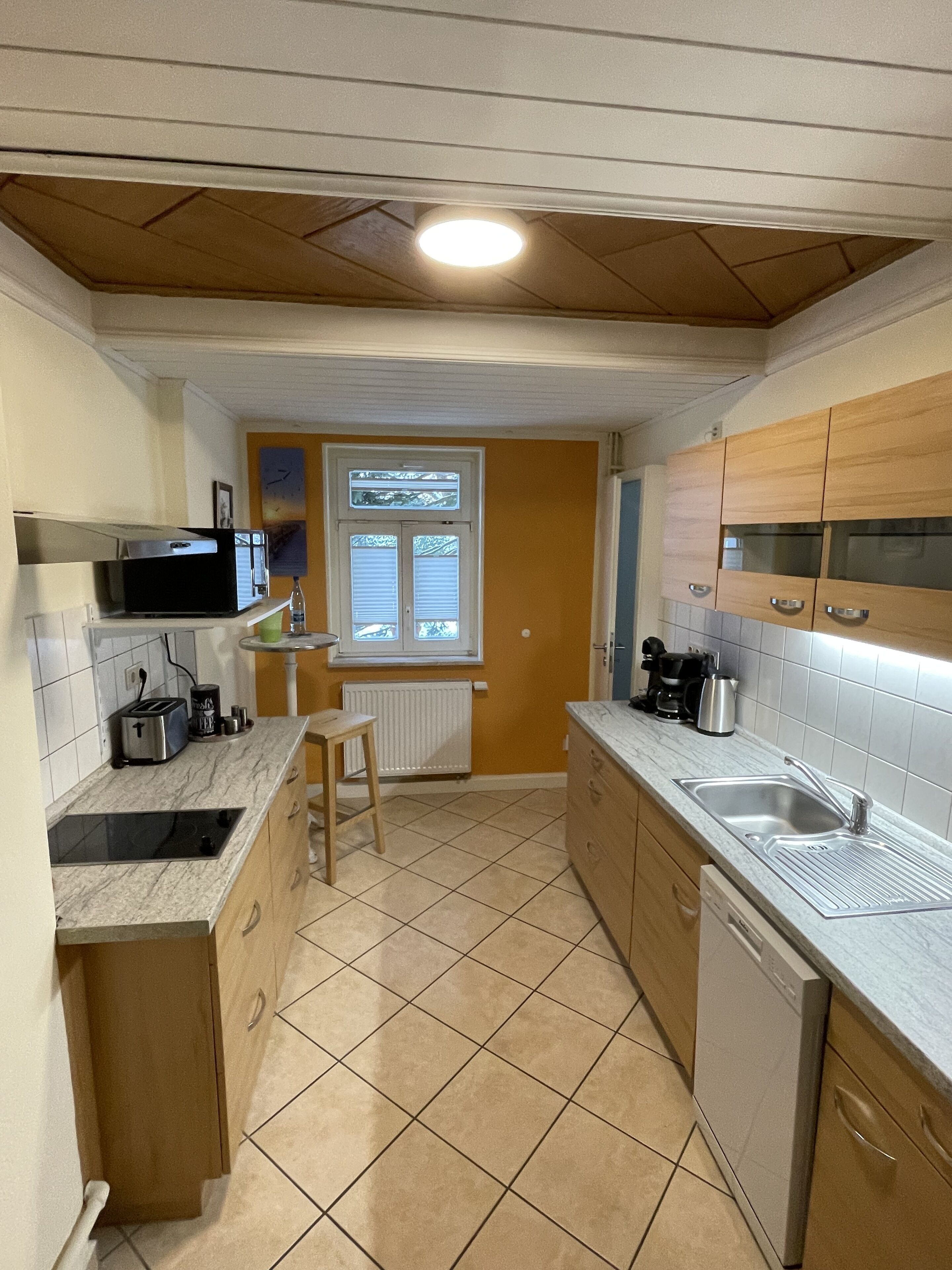 A compact kitchen with a built-in microwave, toaster, and sink. It features wooden cabinets, a tiled floor, and a window with blinds.
