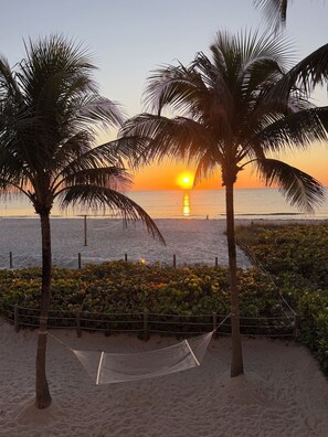 A hammock between palm trees on a sandy beach with a sunset in the background.