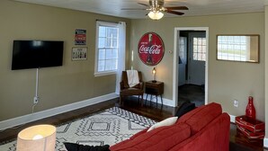 Un salon avec un canapé rouge, un ventilateur de plafond, un téléviseur à écran plat et une enseigne Coca-Cola accrochée au mur.