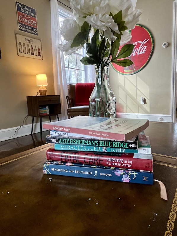 A stack of books on a wooden table, a vase with white flowers, and a Coca-Cola sign on the wall. - 🥤The Coca Cola House 🥤 (Roanoke)
