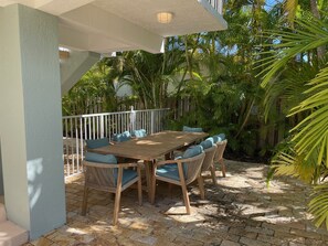 Une terrasse extérieure avec une table en bois et des chaises en osier, entourée d’une végétation luxuriante et d’une clôture blanche.