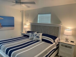Une chambre à coucher bien rangée, avec un lit, deux tables de chevet dotées de lampes, un ventilateur de plafond, une fenêtre avec des stores et un tableau accroché au mur.