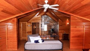 Un dormitorio acogedor en el ático con cama, paredes de madera, ventilador de techo y una zona de cocina visible al fondo.