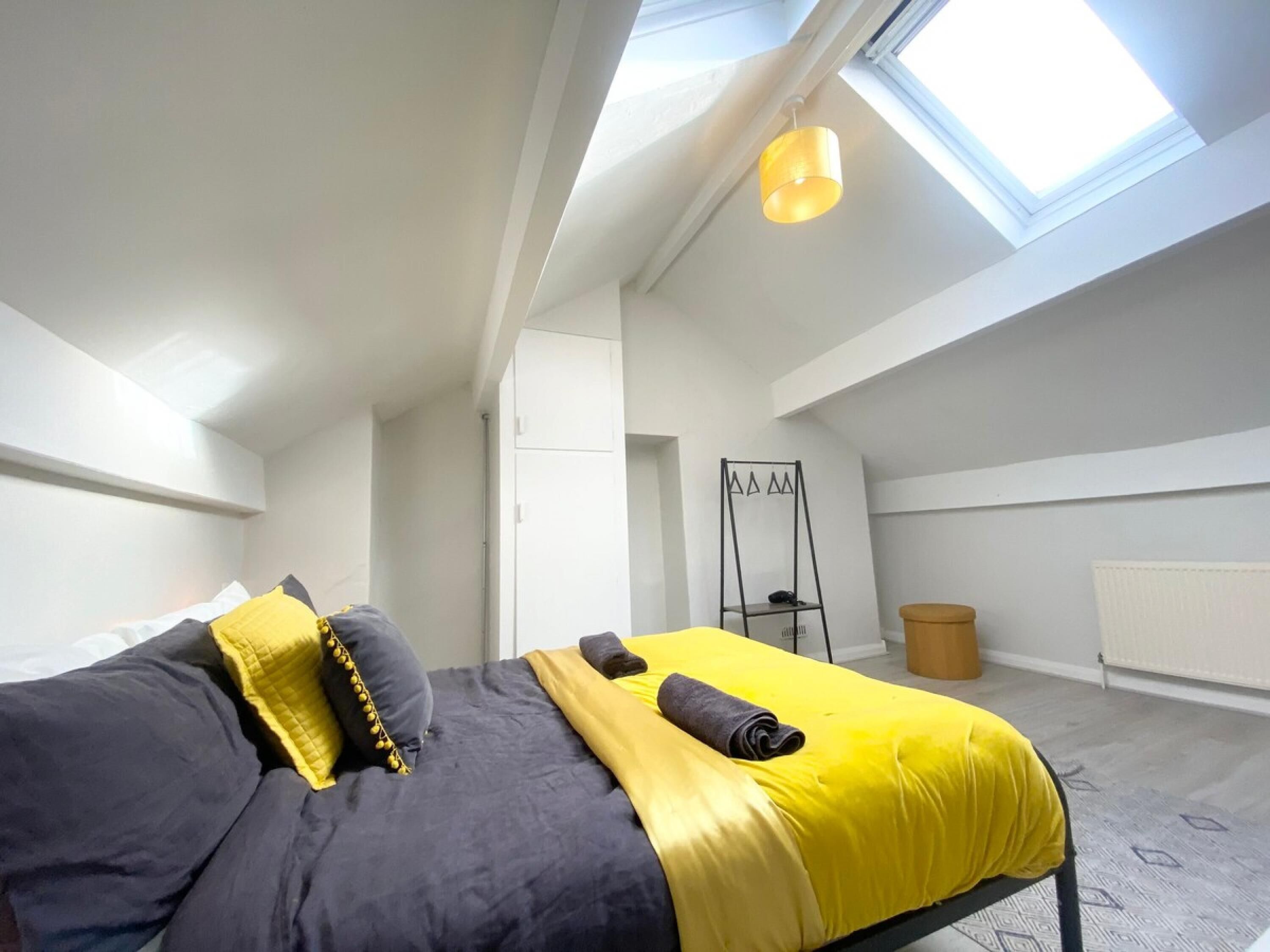 Une chambre à coucher moderne avec une lucarne, un lit avec une literie jaune et grise, un présentoir en métal noir et un radiateur.