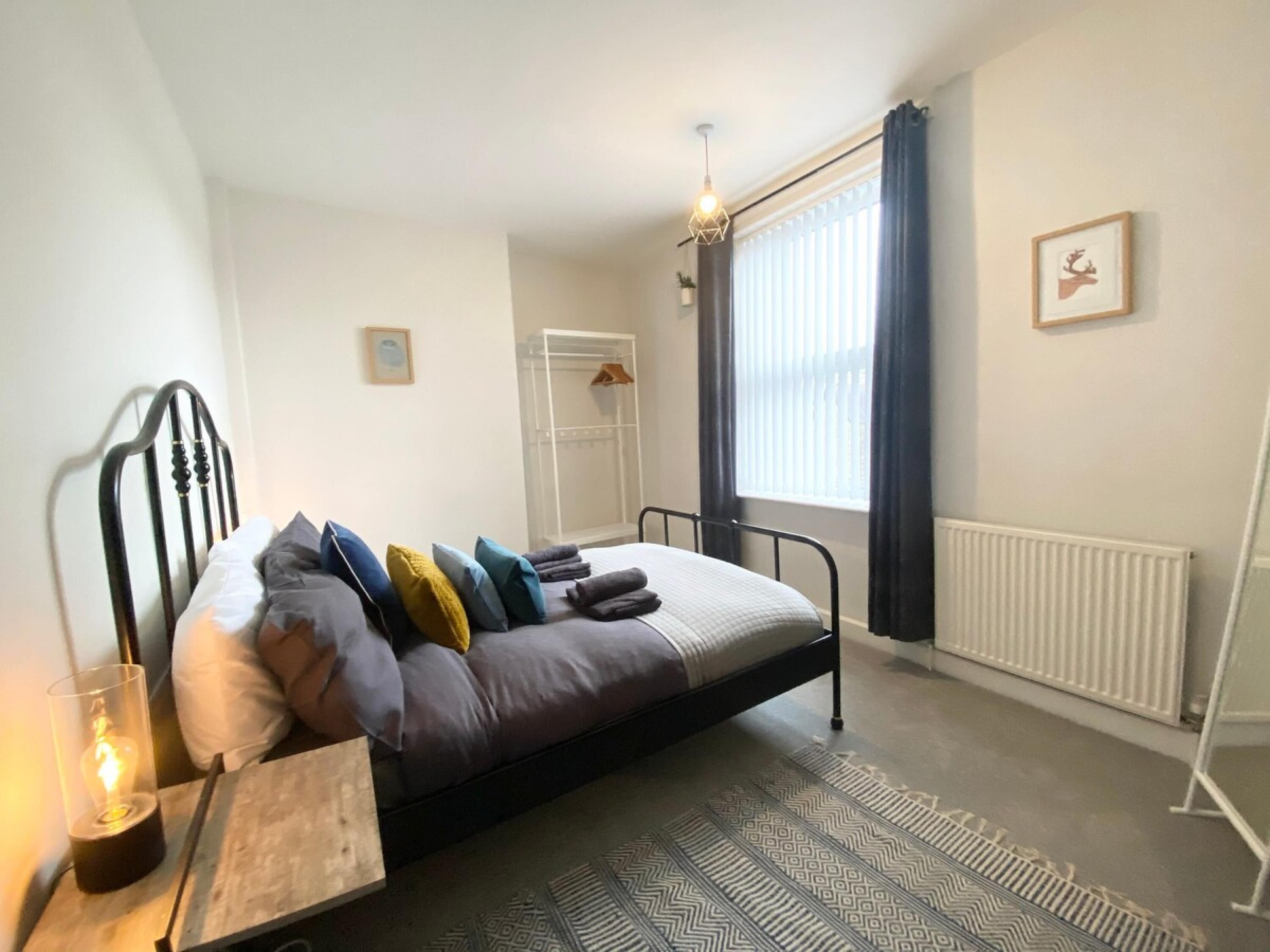 A bedroom with a bed, a bedside table with a lamp, a radiator, a window with curtains, and a framed picture on the wall.