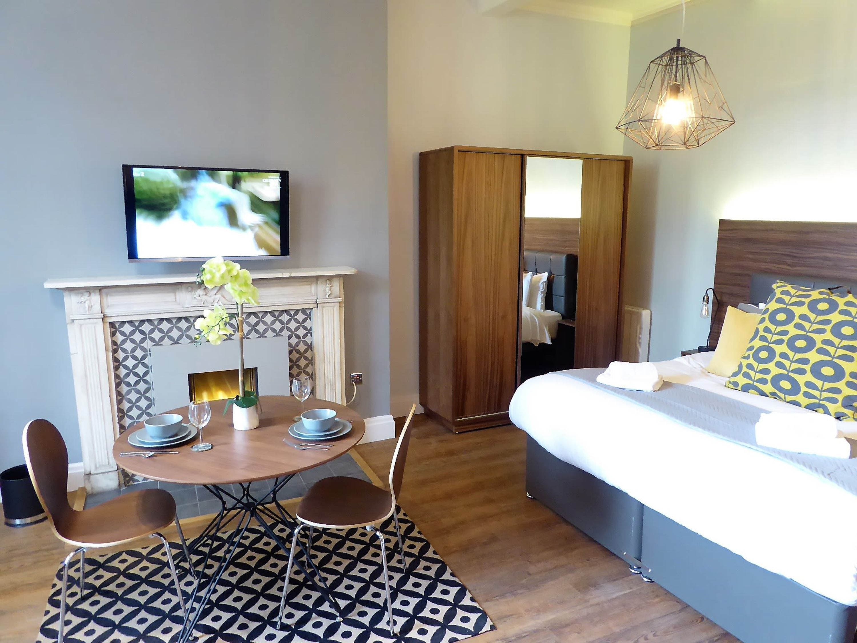 A hotel room with a bed, a TV, a fireplace, a wardrobe, and a dining area with chairs and a table.