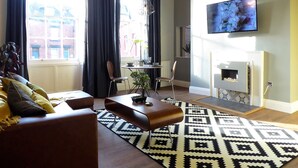 A living room with a sofa, coffee table, and a patterned rug. There is a television mounted on the wall and a small table with a plant. - Claremont The Corson (Leeds)