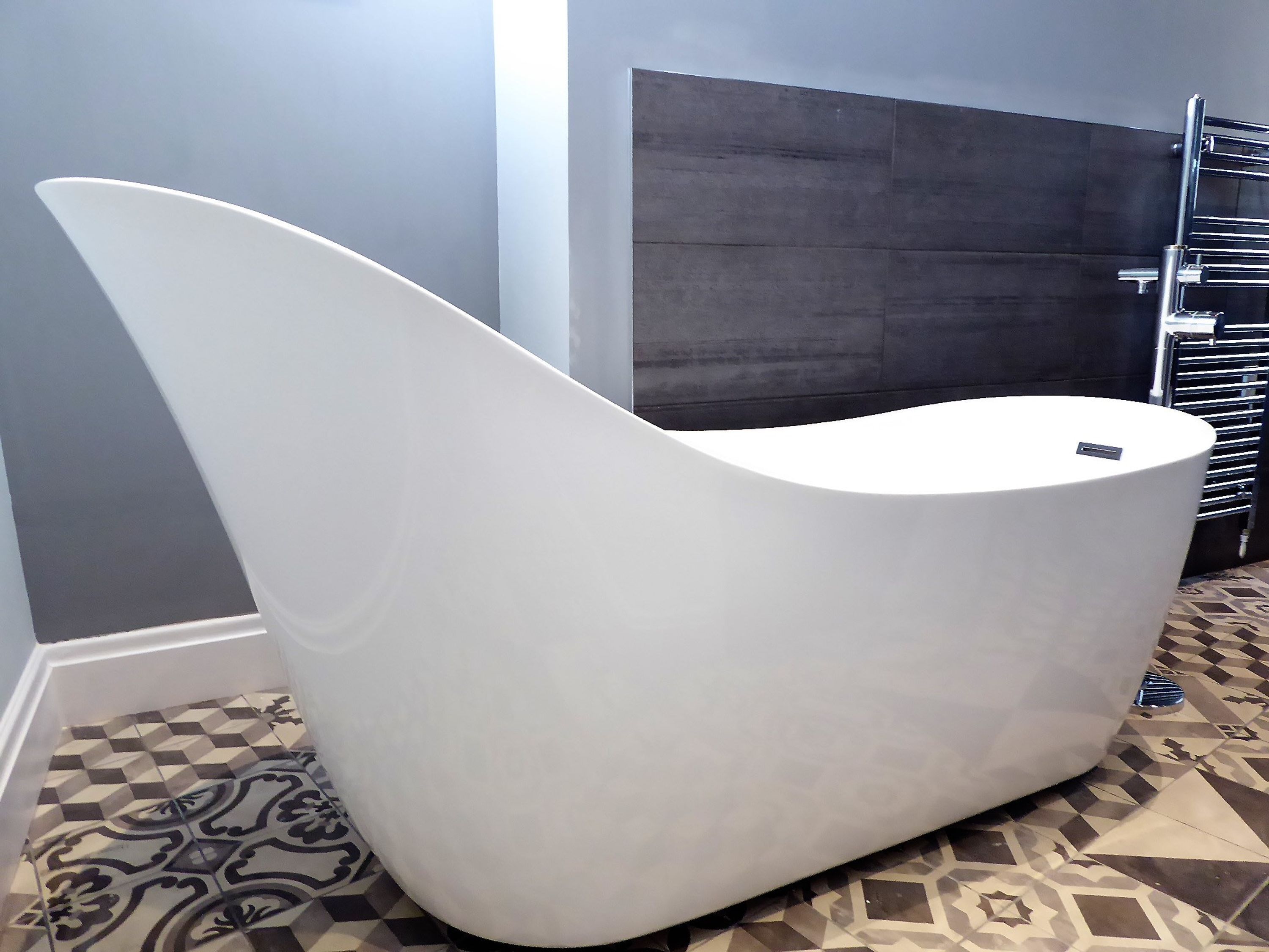 A modern, white, curved bench in a hallway with patterned floor tiles and a dark wall in the background.