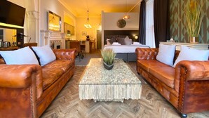 Habitación de hotel con una cama grande, dos sofás de cuero, una mesa de centro y una zona de cocina al fondo.
