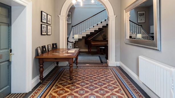 Um corredor com piso estampado, uma mesa de madeira com quadros emoldurados, uma escada e um piano.