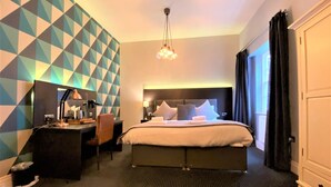 Quarto de hotel com cama, escrivaninha, cadeira, luminária de mesa, espelho, parede com padrão geométrico, luminária pendente e guarda-roupa.