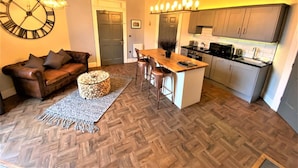 Uma cozinha moderna com uma ilha central, uma sala de jantar com mesa e cadeiras de madeira, e uma sala de estar com sofá e um relógio de parede grande.