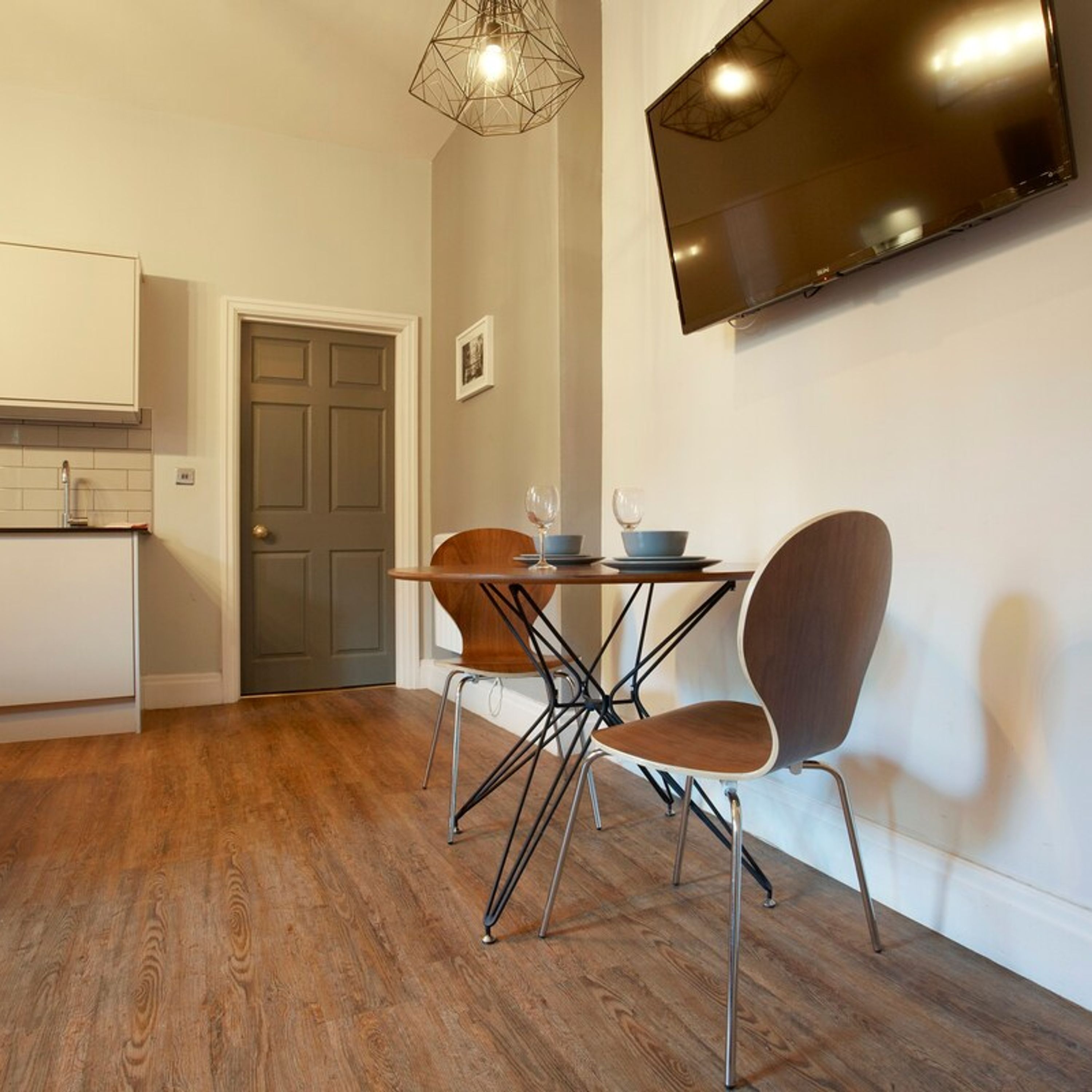 A modern kitchen with wooden flooring, a dining table with chairs, a TV mounted on the wall, and a door leading to another room.