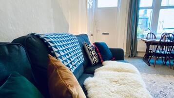 A living room with a dark green sofa, patterned cushions, and a white furry throw. There is a dining area with a table and chairs visible through the window.