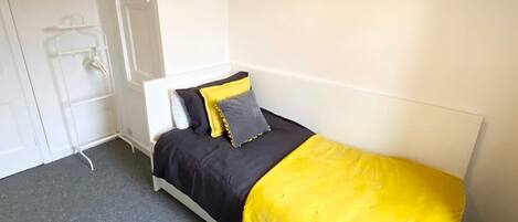 Una habitación pequeña con cama individual, cabecera blanca, alfombra gris y edredón amarillo.