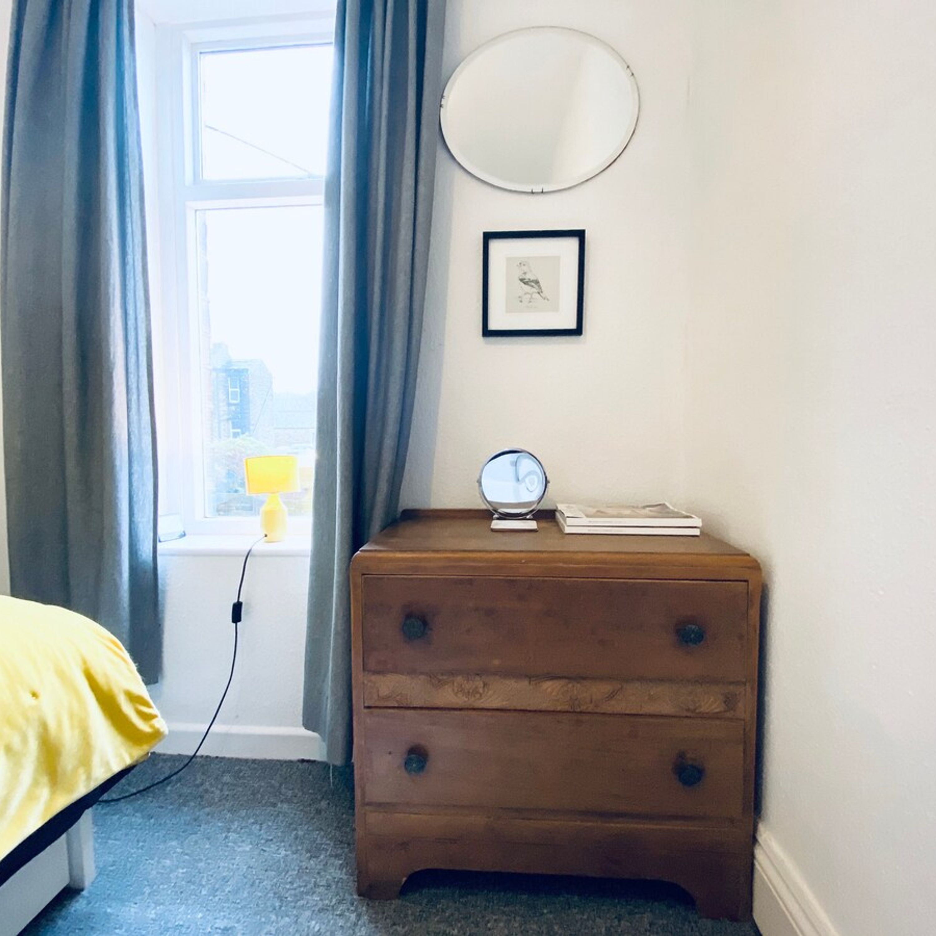 Une chambre avec une commode en bois, un miroir rond et un tableau encadré accroché au mur.