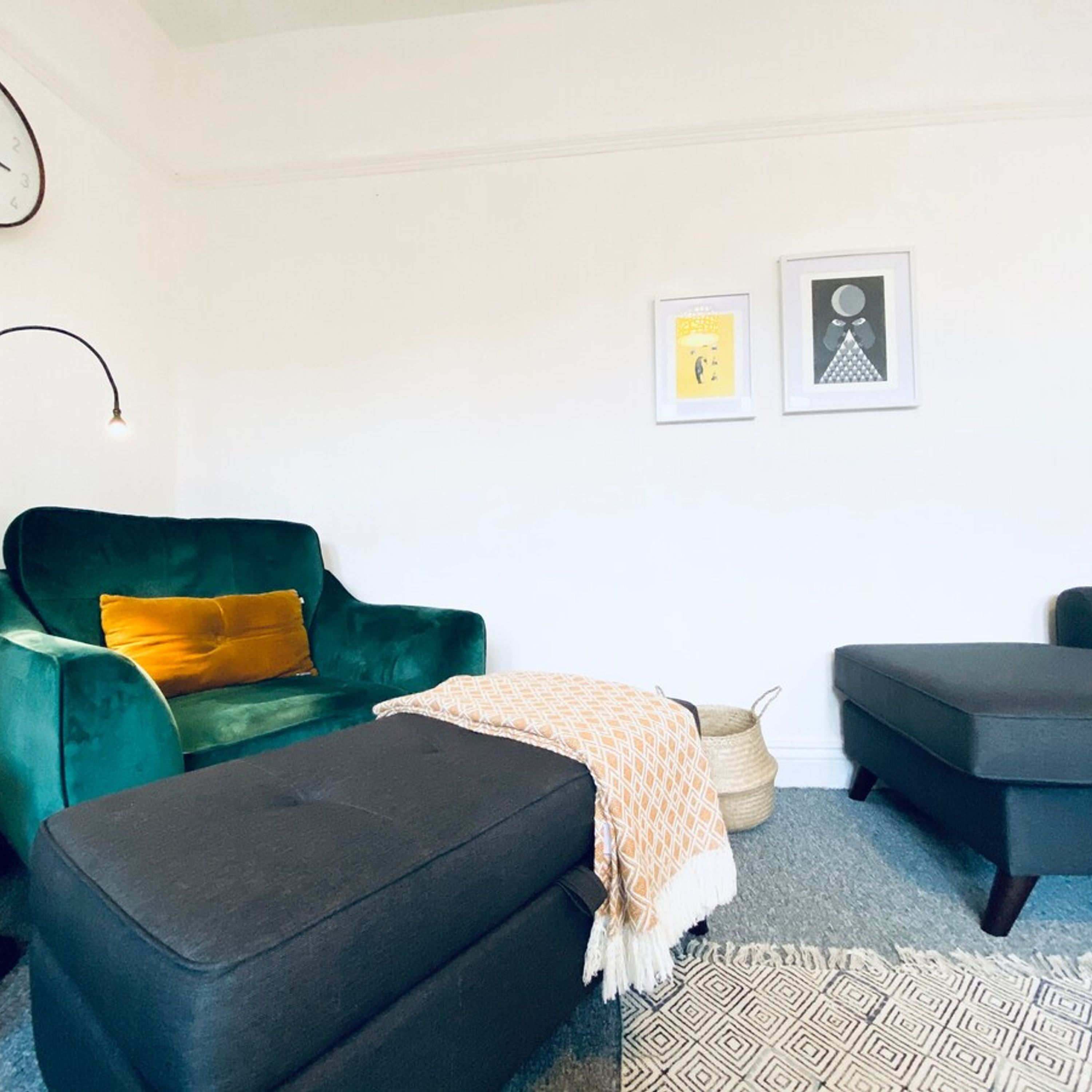 En stue med en grøn sofa, en sort fodskammel og en grå lænestol. To billeder hænger på væggen.