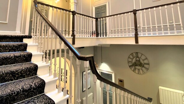 A grand staircase with white balustrades and black handrails, leading to an upper level with a large wall clock. - Claremont The Smeaton (Leeds)