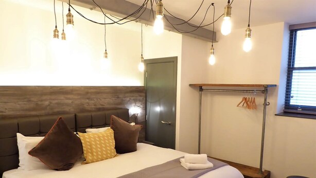 A neatly made bed with a grey and white duvet, two brown pillows, and a white towel. There is a wooden bench with a towel and a hanging rack with clothes. The room has a modern design with exposed brick walls and a window with blinds. - Coach House Stables in Leeds (Leeds)