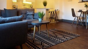 A modern living room with a sofa, coffee table, and a patterned rug. There is a dining area with a table and chairs, and a kitchen in the background. - Coach House Stables in Leeds (Leeds)