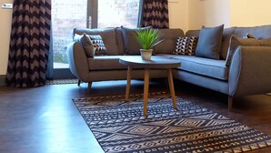 A modern living room with a sectional sofa, a small round table with a plant, patterned curtains, and a view of a brick building outside. - Coach House Stables in Leeds (Leeds)