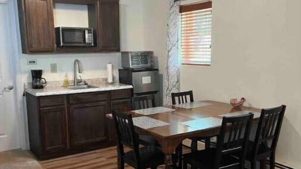 A compact kitchen with dark wooden cabinets, a microwave, and a sink. There is a dining table with four chairs and a rug on the floor.