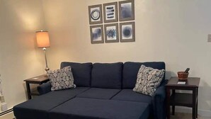 A dark-colored sofa bed with patterned pillows, a side table with a lamp, and a small table with a bowl.